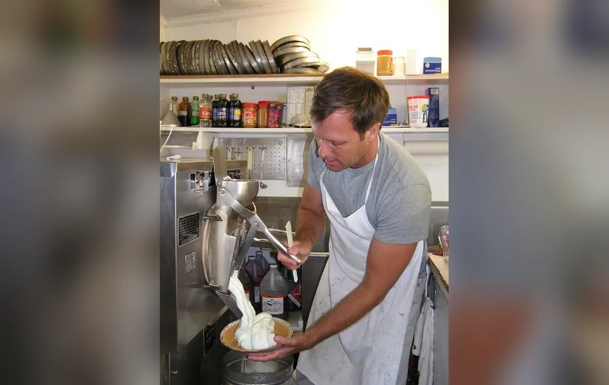 Eric in an apron at Garside's, making ice cream: fresh ice cream pours from the freezer's spout into a pan he holds under it.