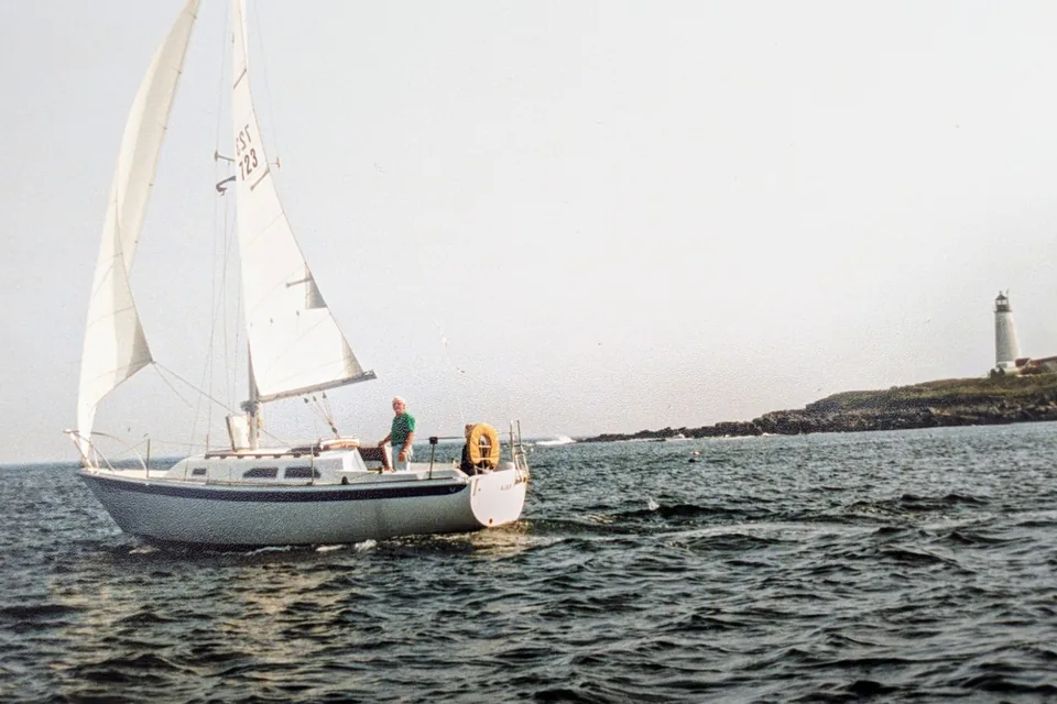 The sailboat Ajax under full sail past a lighthouse on a rocky island, a man in a green shirt at the helm.