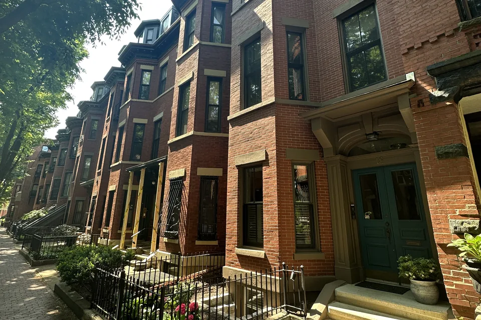 A row of red-brick townhouses with bay windows, a green front door and small iron-fenced gardens on a sunny Boston street.