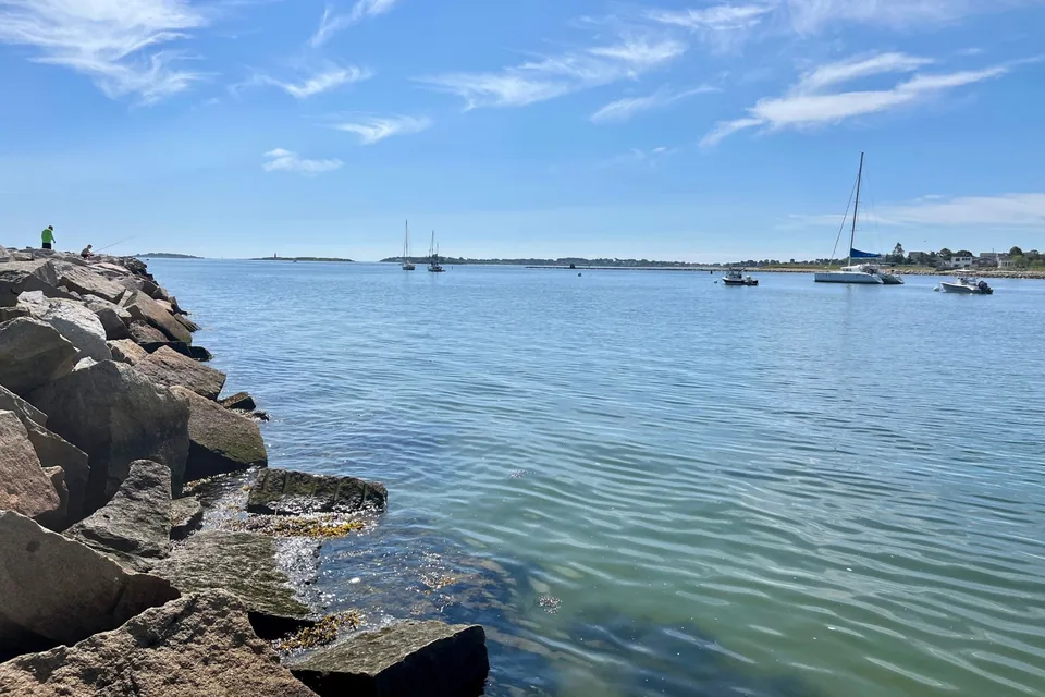 The granite jetty at Camp Ellis on a summer day, sailboats and a catamaran moored in Saco Bay, a lighthouse on an island far off.