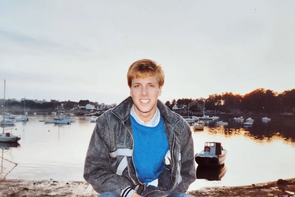Eric as a teenager in a jean jacket, sitting by a harbor in Maine at sunset.