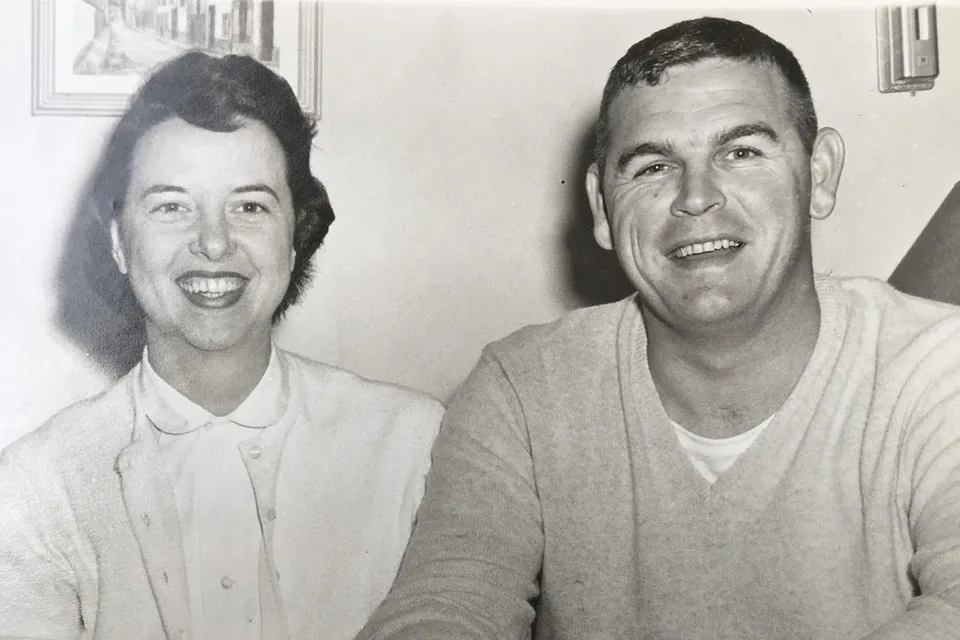 A black-and-white photo of Eric's parents as a young couple, smiling side by side on a sofa, a stack of magazines on the table in front.