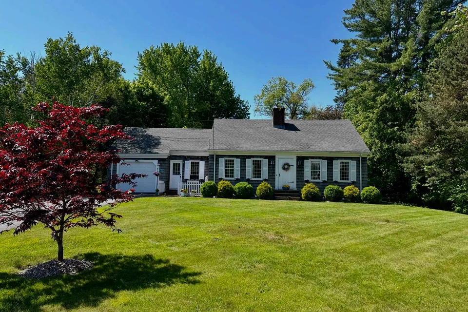 A grey-shingled Cape with white shutters and a red maple on the lawn, under a clear blue sky in Saco.